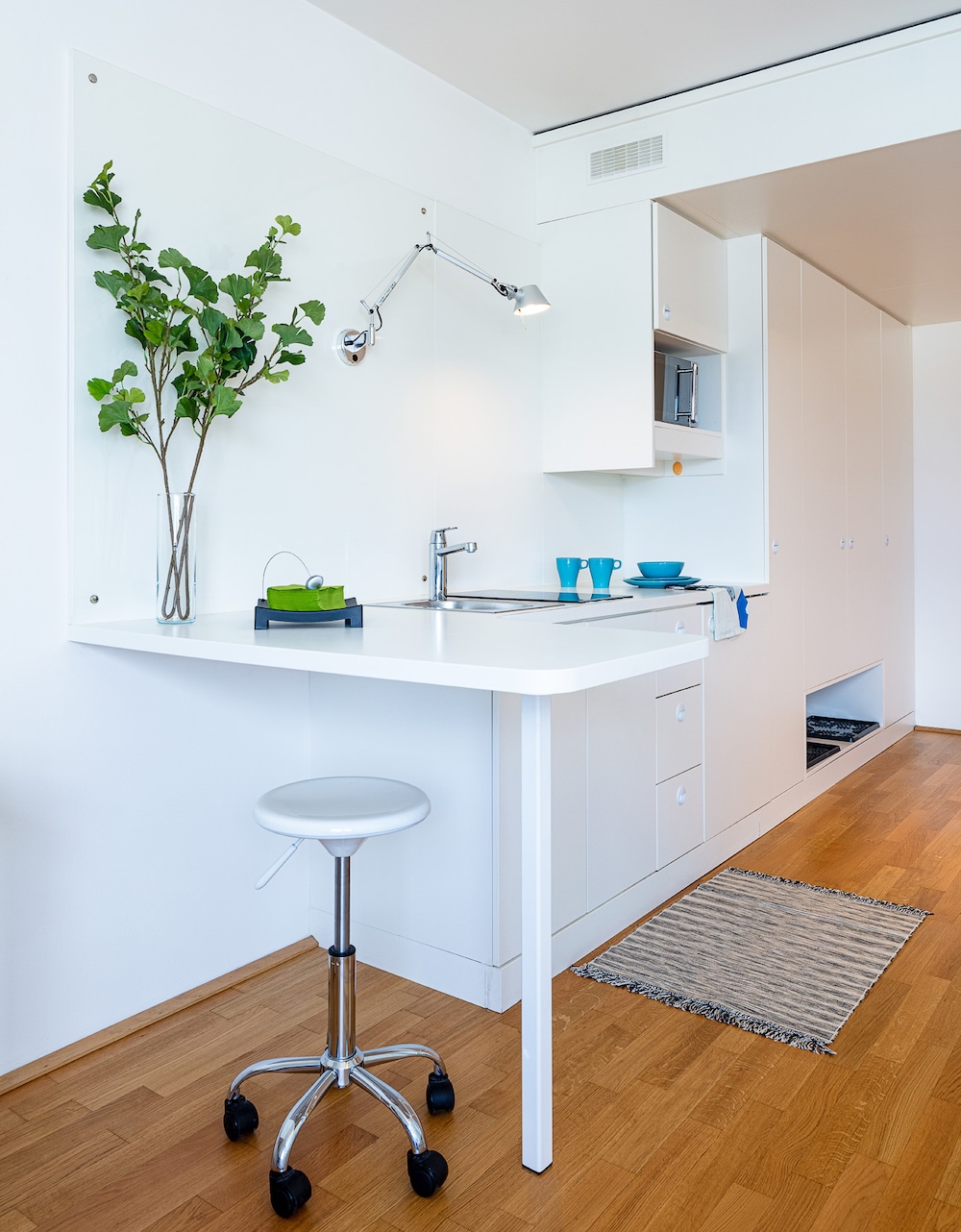 A small white table by the wall with a green plant on top and a stool on a wooden floor in Milestone student housing in Graz.