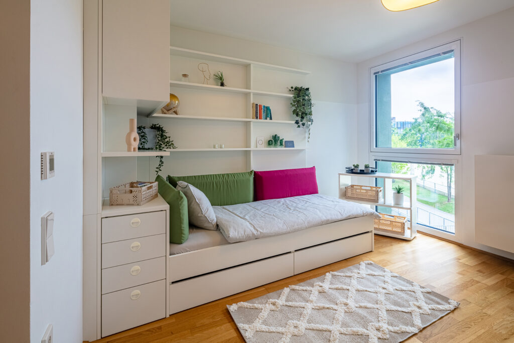 Room of a Milestone by Tribera student dorm in Vienna with a cushioned window seat, built-in drawers, wall shelving and colourful pillows.