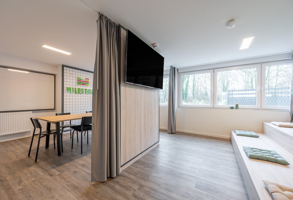 Flexible common room at student housing MILESTONE Bonn with curtains, a wall-mounted television, dining table and tiered seating.