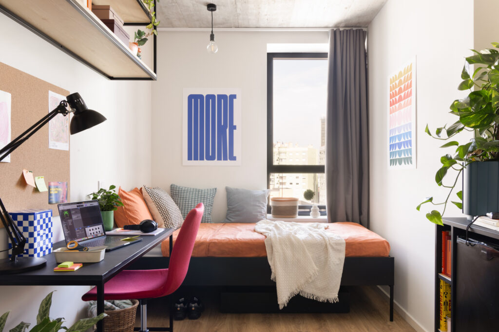Single bed with orange bedding opposite a black study desk and wall artwork in studio room in Milestone Lisbon Olaias student housing.