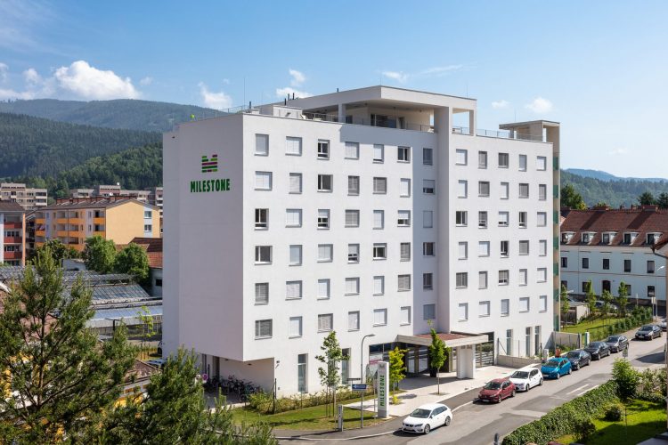 Exterior of MILESTONE Leoben Montan student housing on Schießstattstraße 9, with the white multi-storey residence set against wooded hills.