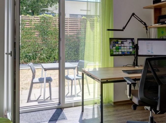 Student room at Milestone Linz with a study desk next to the open terrace door showing an outdoor table with two chairs.