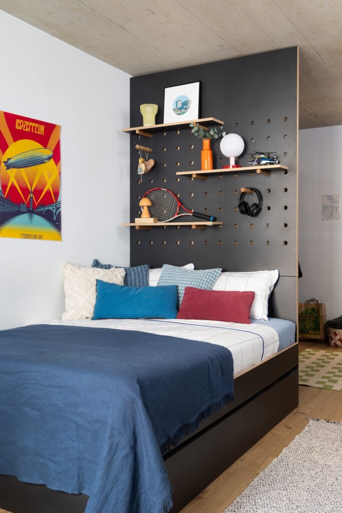 Vienna student residence bed with blue bedding, colourful cushions, pegboard headboard and display shelves above.