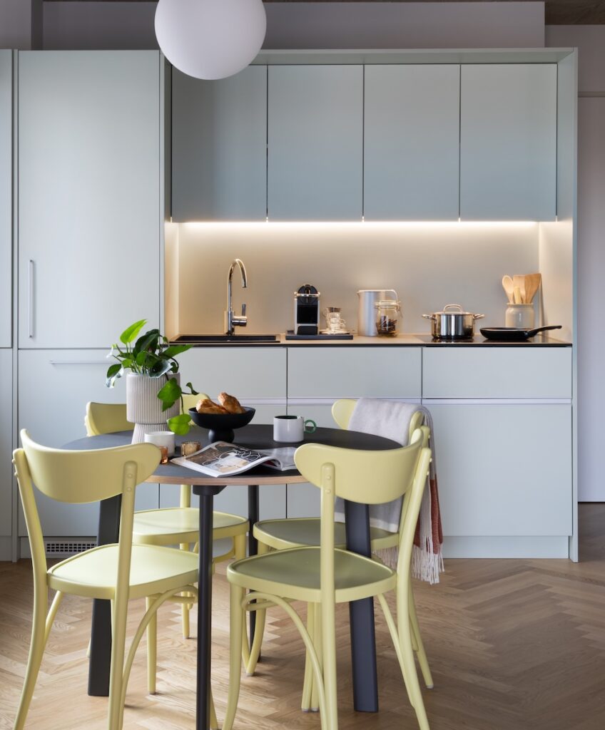 White fitted kitchen and dining table in the Giant Plush Pad student apartment, with under-cabinet lighting and concrete ceiling.