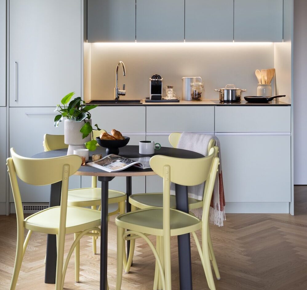 White fitted kitchen and dining table in the Giant Plush Pad student apartment, with under-cabinet lighting and concrete ceiling.