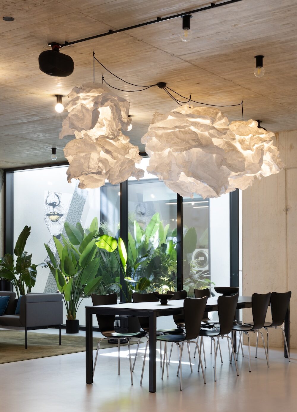 Communal dining area in student residence in Vienna with a long table, black chairs, cloud-like pendant installation and tall indoor plants by the windows.