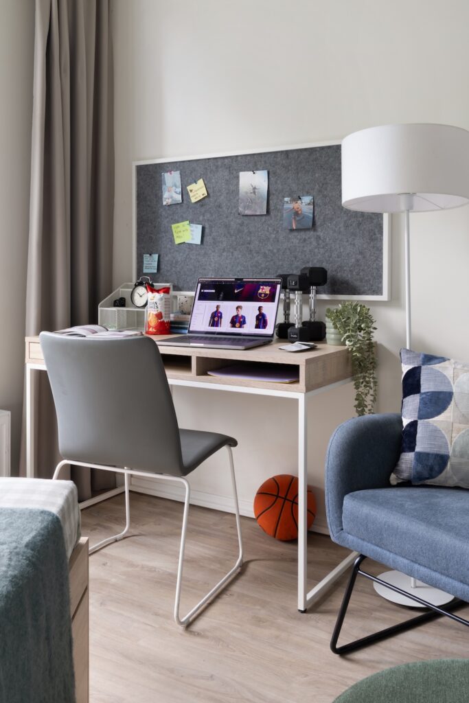 Study desk and pinboard viewed from the sleeping area beside a blue chair.