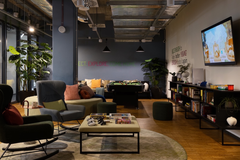 Wide lounge view with mixed seating, bookshelves, plants and a wall-mounted television.