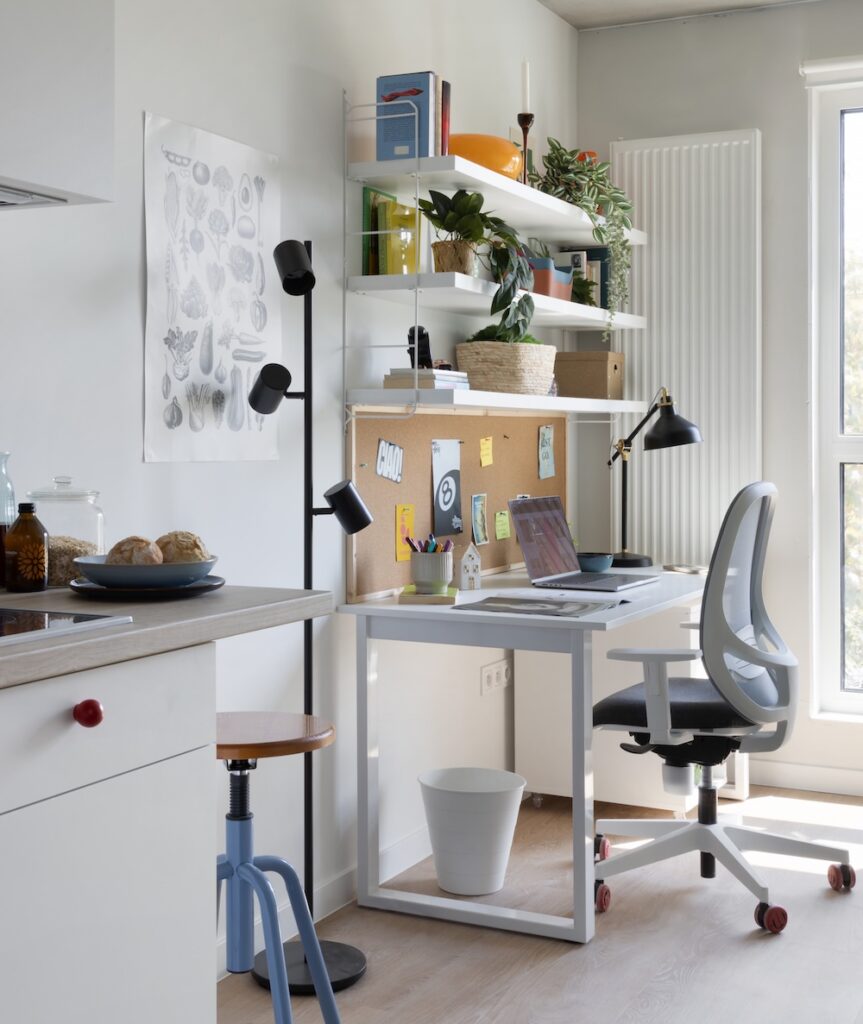 Study desk beside an open kitchenette with shelving, office chair and large window in a student room.