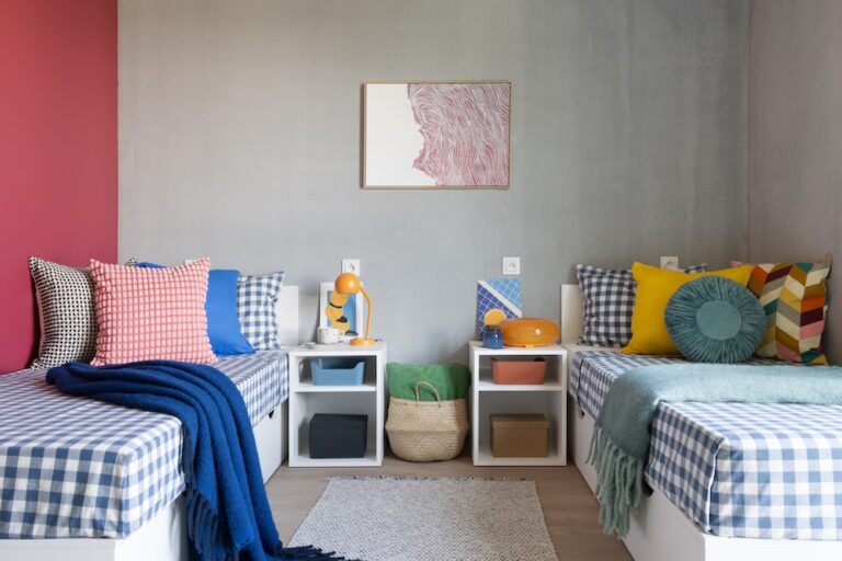 Twin student bedroom with checked bedding, colourful cushions, red accent wall and shared bedside storage.