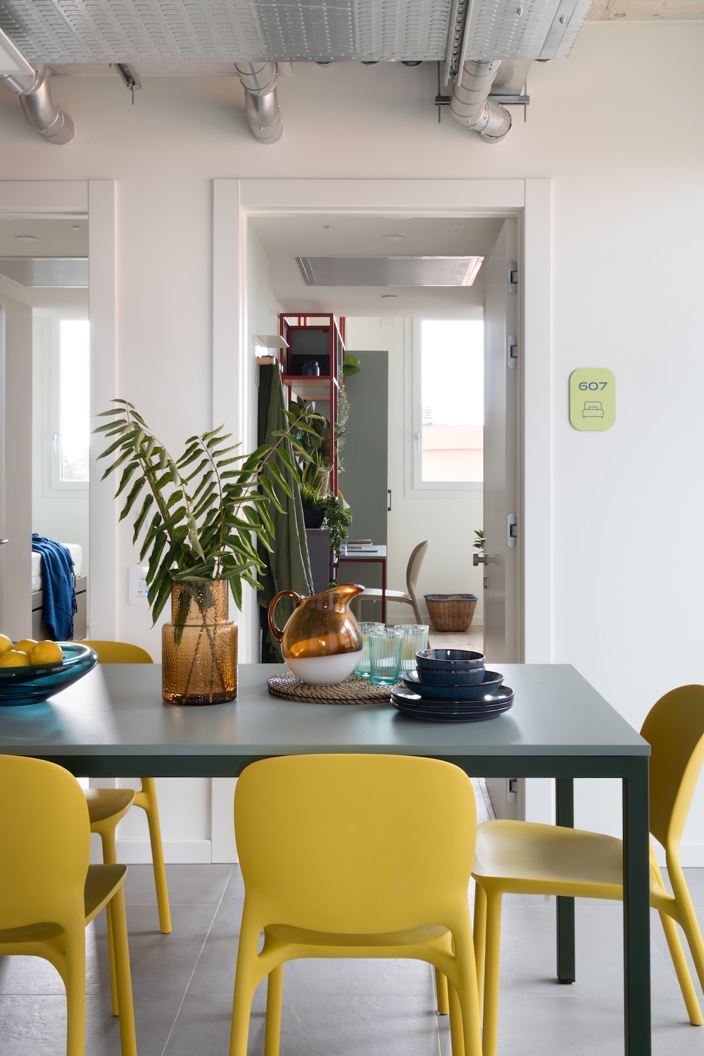 Bright dining space with plants on the table, yellow seating and an open doorway at Tribera student accommodation in Milan.