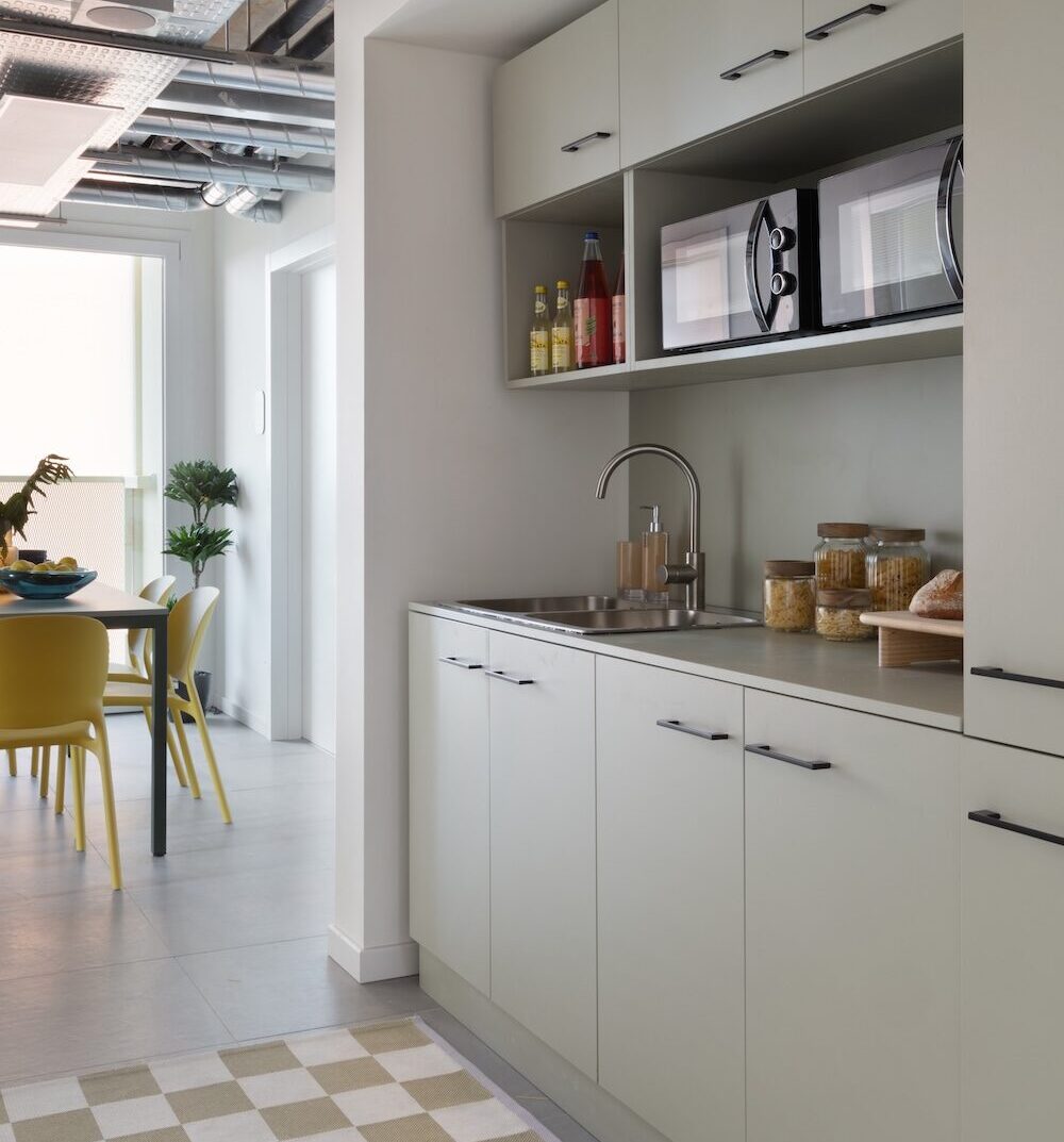Shared kitchen with pale cabinets, chequered floor and dining table with yellow chairs at Tribera student accommodation in Milan.