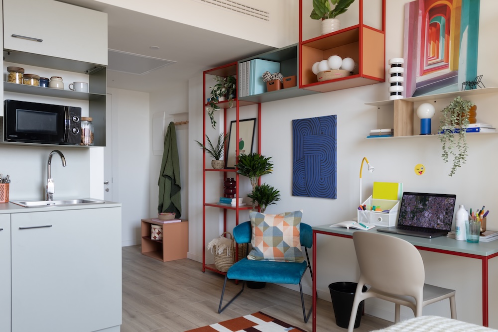 Student room with a study area featuring red shelving, a desk with a white chair and open kitchen area at Tribera student accommodation in Milan.